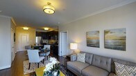 This single-floor residence showcases an open-concept design that integrates the living, dining, and kitchen areas into one cohesive and functional space. The living area is anchored by a gray upholstered sofa accented with chevron-patterned throw pillows, complemented by a yellow coffee table and a patterned area rug that add bold pops of color. Coastal-themed artwork on the neutral walls enhances the relaxed yet polished ambiance. A stylish table lamp provides warm accent lighting alongside a flush-mount ceiling fixture with a gold-toned frame.

The dining area accommodates a compact round table with seating for several, positioned near the kitchen for convenient flow. The kitchen features dark cabinetry, stainless steel appliances including a refrigerator and microwave, and a dark countertop that contrasts cleanly with lighter walls. Crown molding runs throughout the space, adding a refined architectural touch. Dark hardwood-style flooring unifies the entire floor plan, contributing to a warm, contemporary aesthetic. Overall, the space balances practicality with thoughtful design details.