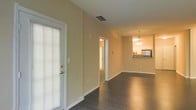 This single-story space offers an open, flowing layout highlighted by dark hardwood flooring that extends throughout the main living areas, providing a sleek and cohesive look underfoot. The walls are finished in a warm, neutral beige tone that complements the flooring and contributes to an inviting ambiance. Vaulted or elevated ceilings add a sense of spaciousness to the interior, while a ceiling-mounted HVAC vent indicates a central air system for year-round comfort.

A door with sidelights and blinds allows natural light to filter into the living area while maintaining privacy. The open-concept design connects the main living space directly to the kitchen, which is visible toward the rear of the unit. The kitchen features a pendant light fixture that hangs above what appears to be a breakfast bar or countertop, creating a defined yet accessible transition between spaces. Stainless steel appliances, including a refrigerator, are visible, adding a modern touch. A hallway leading from the living area suggests access to additional rooms beyond the main living space. Overall, the space presents a clean, functional, and contemporary design aesthetic.