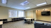 This single-floor multipurpose room is designed to accommodate a variety of meeting and collaborative activities. The space features a drop ceiling with recessed fluorescent panel lighting that ensures consistent, even illumination throughout. Large windows fitted with vertical blinds line one wall, allowing for adjustable natural light to filter into the room.

The layout is thoughtfully arranged to support multiple uses. A central round table surrounded by black stack chairs serves as the primary meeting focal point, encouraging group discussion and collaboration. Along the windowed wall, a long narrow table with additional black chairs provides supplementary workspace, ideal for workshops or supplemental seating. A wall-mounted flat-screen display is positioned at one end of the room, supporting presentations and media viewing. A leather seating piece in the corner adds a more relaxed lounge element to the space.

A set of movable room dividers or folding wall panels along one side suggests the room may be reconfigured or connected to an adjacent area, offering additional flexibility. The patterned carpet flooring adds texture and warmth to the otherwise neutral, professional aesthetic defined by beige and grey tones. Overall, the space presents a clean, functional environment well-suited for meetings, training sessions, or community gatherings.