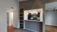 This single-floor residence presents a cohesive interior design anchored by a neutral gray color palette, textured walls, and a combination of wood-look flooring and plush carpeting throughout. The living area is generously sized, featuring vaulted ceilings, a ceiling fan, and a fireplace flanked by built-in open shelving—an ideal focal point for the room. Vertical blinds line a large sliding door and an additional window, allowing natural light to fill the space.

The kitchen is accessible via a pass-through opening framed by a granite-topped breakfast bar, creating a connected yet defined cooking space. Inside, dark espresso cabinetry pairs with granite countertops, a mosaic tile backsplash, and black appliances including a refrigerator and electric range. A skylight above the kitchen adds additional brightness. Built-in shelving units are integrated into the wall adjacent to the kitchen pass-through, offering functional display and storage space.

The bedroom is a quiet, carpeted room with a window fitted with vertical blinds. The bathroom features a dark-framed vanity mirror, granite countertop, undermount sink, and dark wood cabinetry with modern hardware, maintaining the consistent design language seen throughout the residence.