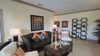 This single-floor living space showcases a thoughtfully designed interior centered around a spacious living area with a warm, contemporary aesthetic. The room features neutral-toned walls that provide a clean backdrop for the carefully curated décor, including framed wall art depicting autumn birch trees alongside smaller complementary pieces that add visual interest and depth.

The living area is anchored by a dark espresso-toned sofa dressed with accent pillows in burnt orange, patterned, and neutral tones, creating a rich, layered look. A matching dark wood coffee table with a lower storage shelf serves as a functional centerpiece, styled with decorative candle holders and reading materials. A light-colored accent chair with striped cushioning provides additional seating and contrast.

A distinctive hexagonal shelving unit along one wall adds a modern, geometric element to the space, displaying decorative objects. Tall, faceted candlestick holders on the coffee table contribute an elegant touch. The carpeted flooring enhances the room's comfort, while recessed lighting and table lamps provide a warm, ambient glow, giving the space an inviting and well-balanced atmosphere.