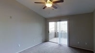 This single-floor space is designed with simplicity and functionality in mind. The main living area features soft, wall-to-wall carpeting that adds warmth and comfort underfoot. A vaulted ceiling rises overhead, creating an open, airy atmosphere and giving the room a generous sense of height. A ceiling fan with an integrated light fixture is centrally mounted, providing both air circulation and ambient lighting.

Neutral gray walls offer a clean, versatile backdrop suited to a variety of interior styles. A sliding glass door, dressed with vertical blinds, serves as both a source of natural light and a direct access point to a private outdoor balcony. The balcony extends the living space outdoors and features a railing, allowing for open views of the surrounding area. Natural light filters through the glass door, brightening the interior during daylight hours.

The overall ambiance is calm and understated, with a practical layout that balances indoor comfort with outdoor accessibility. The textured ceiling and simple architectural details contribute to a straightforward, livable design well-suited for everyday use.
