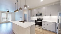This single-floor residence offers a well-designed layout across two bedrooms and two bathrooms, thoughtfully finished with contemporary materials throughout. The open-concept kitchen and living area serves as the heart of the home, anchored by a large waterfall-edge quartz island with an undermount sink and matte black faucet. Gray flat-front cabinetry lines the kitchen walls, complemented by a white subway tile backsplash, under-cabinet lighting, and a full suite of stainless steel appliances including a french door refrigerator, electric range, and built-in microwave. Pendant lighting clusters above the island add a modern design touch. Light-toned hardwood flooring runs through the main living areas, while plush carpet covers the bedrooms. The primary bedroom features a ceiling fan and direct access to a well-appointed bathroom with quartz countertops, gray cabinetry with black hardware, wood-look tile flooring, and a soaking tub with large-format tile surround. The secondary bedroom includes a ceiling fan, natural light from a window, and closet storage. Both closets feature built-in shelving and hanging rods. The living area includes French doors that introduce natural light and an open, airy ambiance throughout.