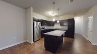 This single-floor residence offers a thoughtfully designed layout with modern finishes throughout. The kitchen serves as a standout feature, showcasing dark espresso cabinetry, granite-style countertops, a full suite of stainless steel appliances including a refrigerator, range, microwave, and dishwasher, and a central island with additional counter space. Pendant lighting and a built-in wine rack add functional elegance to the space. Rich hardwood-style flooring extends through the kitchen and open living areas, creating a warm and cohesive aesthetic. The spacious living area flows seamlessly from the kitchen, offering an open and versatile layout. The bedrooms are finished with plush carpeting, ceiling fans, and window blinds that allow for natural light while maintaining privacy. Each bedroom includes access to generous closet space, with at least one walk-in closet featuring built-in shelving and hanging rods. The bathrooms are finished with matching dark vanity cabinetry, granite-style countertops, framed mirrors, and a shower-tub combination. Consistent hardwood-style flooring ties the bathrooms to the rest of the home's design language. An attached garage with an automatic opener provides additional storage and secure parking. The overall ambiance is clean, contemporary, and move-in ready.