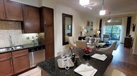 This refined single-floor residence offers an open-concept design that seamlessly connects the kitchen, dining, and living areas into one cohesive and elegant living space. The kitchen is appointed with rich dark wood cabinetry, gleaming dark granite countertops with subtle veining, stainless steel appliances including a dishwasher, and under-cabinet accent lighting that adds warmth to the space. A large kitchen island with the same dark granite surface serves as both a prep area and a casual dining spot, complete with place settings and stemware on display. A large framed mirror in a dark wood surround is positioned near the dining area, enhancing the sense of depth and light throughout. The dining area features sophisticated upholstered chairs with a glass-top table, while curated wall art adds a gallery-like quality to the surrounding walls. The adjacent living area is warmly lit with floor lamps and benefits from natural light filtering in through large windows draped with elegant curtains. Track lighting and a pendant light illuminate the kitchen and dining zones, creating a layered lighting scheme. The overall ambiance is polished and inviting, with a consistent palette of warm neutrals, deep wood tones, and refined finishes throughout.
