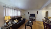 This single-floor residence offers a practical and comfortable layout with a cohesive design throughout. The living room is an inviting space anchored by a dark leather sofa, light hardwood-style flooring, and large windows dressed with sheer curtains that allow natural light to filter in. A dedicated media area with a wall-mounted TV and open shelving completes the room.

The dining area flows naturally from the living space, featuring a glass-top dining table with seating for six, and connects seamlessly to the kitchen via a partial partition with counter seating. The kitchen is well-equipped with dark espresso cabinetry, granite-style countertops, a gas range, and tile flooring, providing ample storage and prep space.

The residence includes multiple bedrooms, each carpeted and outfitted with windows that bring in soft natural light. The bedrooms offer closet storage and enough space to comfortably accommodate dressers and additional furnishings.

The bathroom retains a classic character with its distinctive blue ceramic wall tile, mosaic tile flooring, and a full bathtub with shower. Throughout the space, neutral wall tones provide a clean, understated backdrop that ties each room together into a cohesive and functional home.
