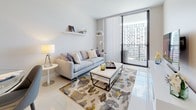 This single-floor residence showcases a contemporary design characterized by clean lines, neutral tones, and large-format polished tile flooring throughout. The open-concept living area is bright and welcoming, anchored by floor-to-ceiling sliding glass doors that lead to a private balcony with urban views. Floating wall shelves, a light gray sofa, and a vibrant patterned area rug add personality to the space.

The kitchen is thoughtfully designed with wood-grain cabinetry, granite countertops, a mosaic tile backsplash, and a full suite of stainless steel appliances. A central island with bar seating provides additional prep space and casual dining. Track lighting overhead ensures excellent illumination throughout.

The primary bedroom features a soft blue accent wall, an upholstered panel headboard, and matching white nightstands with contemporary lamps. The en-suite bathroom includes granite countertops, wood-toned vanity cabinetry, a walk-in closet, and polished tile flooring. A second bedroom benefits from a large window that draws in natural light, complemented by coordinating furnishings and artwork. A second full bathroom with a soaking tub and tiled surround serves the remaining rooms.