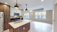 Located at 2432 Albany Ave in West Hartford, CT, this contemporary single-floor unit offers a thoughtfully designed open-concept layout. The kitchen serves as a central highlight, featuring a large island with a white quartz countertop, an undermount sink, and a wood-grain base that adds warmth to the modern aesthetic. White upper cabinetry is complemented by rich walnut-toned lower cabinets and tall pantry storage, while stainless steel appliances — including a refrigerator, electric range, and built-in microwave — complete the space. Stylish pendant lights hang above the island, providing both task lighting and visual appeal. The kitchen flows seamlessly into the living area, which is bathed in natural light through multiple large windows fitted with roller shades, offering views of the surrounding neighborhood. Light hardwood-style flooring runs throughout the unit, enhancing the bright, airy atmosphere. The bathroom is finished with a wood-grain vanity, white quartz countertop, a large wall mirror, and a tiled walk-in shower, maintaining the clean, modern character found throughout the unit. An additional room provides flexible space with its own window and the same consistent flooring and neutral wall tones.