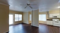 This single-floor residence offers a thoughtfully designed layout that balances open living space with practical storage solutions. The main living area is a standout, featuring rich, dark hardwood flooring that extends throughout and creates a warm, inviting atmosphere. Crown molding adds a refined architectural detail along the upper walls, while warm golden-toned paint contributes to the space's welcoming ambiance. A ceiling fan with a light fixture serves as both a functional and decorative element overhead. Multiple windows dressed with vertical blinds flood the area with natural light, creating a bright and airy feel. A partial column divides the living space, subtly defining separate functional zones while maintaining an open-concept flow. Track lighting in the adjacent area provides additional illumination options. Transitioning from the main living area, a hallway leads to a dedicated built-in shelving alcove, offering generous vertical storage across multiple shelves — ideal for organization and display. The flooring shifts to light-toned tile in this transitional zone, providing a clean visual contrast. Overall, the space combines classic design touches with practical features to deliver a comfortable and functional single-floor living environment.