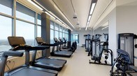 This contemporary fitness center offers a well-appointed, single-floor exercise facility designed to support a full range of workout routines. The space is defined by its expansive floor-to-ceiling windows that run along one side of the room, flooding the interior with natural light and offering sweeping panoramic views of the surrounding cityscape — a motivating backdrop for any workout.

The cardio zone is lined with a row of treadmills and elliptical machines, all positioned to take full advantage of the stunning views. Adjacent to the cardio area, a dedicated strength training section houses a variety of cable machines and resistance equipment, providing users with comprehensive options for muscle conditioning and functional fitness.

The high ceilings contribute to an open, airy atmosphere, while recessed linear LED lighting ensures the space remains well-lit throughout. Large mirrors are mounted along one wall, enhancing the sense of spaciousness and allowing users to monitor their form during exercise.

The neutral color palette — white walls, grey flooring, and dark equipment — gives the facility a clean, professional aesthetic. The thoughtful layout ensures clear pathways between equipment, promoting a safe and comfortable training environment. Overall, this fitness center combines modern design with practical functionality.
