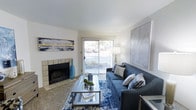 This single-floor space centers around a well-appointed living room that balances comfort and style. A tile-surround fireplace serves as a natural focal point along one wall, adding warmth and architectural character to the room. Adjacent to the fireplace, a sliding glass door floods the space with natural light while providing direct access to an outdoor area, offering views of the surrounding greenery.

The living room is generously sized, accommodating a full sofa and coffee table arrangement with ease. A coordinated blue color palette runs throughout the furnishings, from the navy sofa accented with mixed throw pillows to a boldly patterned blue area rug anchoring the seating area. A glass-top coffee table adds a light, airy touch that keeps the space feeling open.

The walls are painted a clean, crisp white, enhancing the brightness of the room. Large framed artwork on opposing walls adds visual interest and reinforces the contemporary aesthetic. Carpeted flooring contributes a soft, comfortable underfoot feel. Overall, the space presents a cohesive, inviting atmosphere with thoughtful design details throughout.