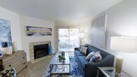 This single-floor space centers around a well-appointed living room that balances comfort and style. A tile-surround fireplace serves as a natural focal point along one wall, adding warmth and architectural character to the room. Adjacent to the fireplace, a sliding glass door floods the space with natural light while providing direct access to an outdoor area, offering views of the surrounding greenery.

The living room is generously sized, accommodating a full sofa and coffee table arrangement with ease. A coordinated blue color palette runs throughout the furnishings, from the navy sofa accented with mixed throw pillows to a boldly patterned blue area rug anchoring the seating area. A glass-top coffee table adds a light, airy touch that keeps the space feeling open.

The walls are painted a clean, crisp white, enhancing the brightness of the room. Large framed artwork on opposing walls adds visual interest and reinforces the contemporary aesthetic. Carpeted flooring contributes a soft, comfortable underfoot feel. Overall, the space presents a cohesive, inviting atmosphere with thoughtful design details throughout.