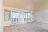 This single-floor residence offers a thoughtfully laid-out interior that balances open living space with practical functionality. The main living area is generously proportioned, featuring wall-to-wall carpet, smooth ceilings, and an abundance of natural light streaming through large windows dressed with vertical blinds. A sliding glass door leads to an outdoor balcony or patio, framing views of lush, mature greenery that bring a natural, peaceful ambiance to the interior. An adjacent carpeted room, visible through an open passage, extends the living space further and shares the same bright, airy character.

The galley-style kitchen is efficiently designed with rich, warm-toned wood cabinetry lining both walls from floor to ceiling, providing ample storage. A double sink, white dishwasher, electric range, and refrigerator are all included, with light-toned countertops and vinyl tile flooring completing the clean, functional aesthetic.

The bathroom features a full soaking tub with a tub surround, a vanity with wood cabinetry, and tile flooring, offering a simple and clean space. Throughout the residence, wood-panel interior doors add a warm, classic touch. The overall layout, as reflected in the dollhouse view, presents a compact yet well-organized floor plan suited for efficient, comfortable living.