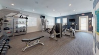 Located at 1100 Cannonball Run in Knightdale, NC, this single-floor fitness center is thoughtfully designed to support a comprehensive workout experience. The space is organized into distinct functional zones, each outfitted with professional-grade equipment. Bold navy blue accent walls contrast with crisp white ceilings and trim, creating an energizing atmosphere throughout.

The cardio zone features a row of TRUE treadmills and elliptical machines, positioned alongside windows with white blinds that allow natural light to fill the room. A wall-mounted TV keeps members entertained during workouts. The strength training area is equipped with a multi-functional cable machine, a leg press/extension machine, and an adjustable weight bench, providing a full range of resistance training options. A dedicated free weights section includes a tiered dumbbell rack flanked by floor-to-ceiling mirrors that enhance the sense of space.

An additional area, separated by sliding glass doors, appears to serve as a studio or stretching space, offering a quieter zone for flexibility work or group fitness. The flooring throughout consists of durable, textured carpet tile, and ceiling fans and recessed lighting ensure the environment remains comfortable and well-lit. The overall layout is spacious, functional, and well-maintained, making it a well-rounded amenity facility.