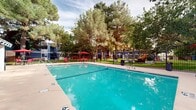 This outdoor amenity area centers around a large, sparkling swimming pool with clearly marked depth indicators, accommodating both casual swimmers and lap swimmers. The pool's vivid turquoise water is complemented by a wide concrete deck that surrounds the perimeter, providing ample space for movement and relaxation. Multiple red lounge chairs and umbrella-shaded patio tables are positioned around the deck, offering designated areas for sunbathing and socializing. A single-story clubhouse building with a covered porch and columned entryway is situated directly adjacent to the pool, serving as a functional hub for the recreational area. The structure features a neutral stucco exterior consistent with a Southwestern architectural style. The entire pool area is enclosed by a black wrought iron fence, ensuring safety and defined boundaries. Beyond the fence, well-maintained green lawn areas extend outward, bordered by tall, mature pine and deciduous trees that provide natural shade and a sense of privacy. A children's playground with colorful equipment and a covered pergola structure are visible within the secured grounds, adding family-friendly functionality. The overall environment is open, airy, and inviting.