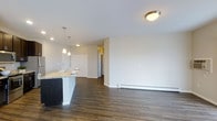 This single-floor space showcases a modern, open-concept design that integrates the kitchen and living areas into one cohesive layout. The kitchen features a prominent island topped with granite-style countertops, a stainless steel sink, and elegant pendant lighting that hangs above, providing both functional task lighting and a stylish focal point. The cabinetry is finished in a crisp white, complementing the clean, neutral palette throughout the space.

Dark hardwood flooring extends across the entire open area, adding warmth and contrast to the bright white walls and ceilings. Flush-mount ceiling fixtures provide ambient lighting, ensuring the space feels well-lit throughout. A baseboard heating unit runs along the far wall, indicating a practical and efficient heating solution.

The living area is generously sized, offering ample room for a variety of furniture arrangements. A hallway visible in the background suggests additional rooms beyond the open living space, providing a sense of depth and privacy. Overall, the space presents a clean, contemporary aesthetic with a functional, well-considered layout.