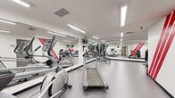 This fitness facility at 2470 Southvale Crescent in Ottawa, Ontario offers a compact yet comprehensively equipped workout space designed to support a full range of exercise routines. The room features a clean, modern interior with white walls accented by bold red diagonal stripes, giving the space an energetic and motivating aesthetic. Large floor-to-ceiling mirrors line one wall, visually expanding the room and allowing users to monitor their form during workouts.

The cardio section is well-stocked with treadmills, elliptical trainers, and a stationary bike, each positioned to make efficient use of the available floor space. A mounted flat-screen television provides entertainment during cardio sessions. The strength and free weights area includes a full rack of dumbbells, a weight bench, a rowing machine, and an assortment of exercise balls in various sizes, catering to both strength training and flexibility work.

Bright, recessed linear fluorescent lighting runs across the ceiling, ensuring the entire space is evenly and well-lit. The durable, smooth flooring is practical and easy to maintain. A motivational quote displayed on the wall near the entrance adds an encouraging touch. The facility is accessed through a key-controlled glass door and includes a wall clock, a waste bin, and posted informational materials, reflecting a thoughtfully organized and functional fitness environment.
