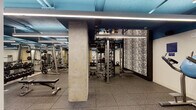 This fitness center is a fully outfitted, single-floor gym designed to support a wide range of workout routines. The space features an industrial aesthetic, with exposed ductwork and structural columns painted in a striking deep blue, complemented by sleek linear LED lighting that runs across the ceiling to provide consistent, bright illumination throughout. The flooring throughout the gym consists of durable rubber tiles, offering both safety and comfort during exercise. The strength training area is well-appointed with a variety of machines, including cable systems, Smith machines, multi-function racks, and a full dumbbell rack with accompanying adjustable benches. A standout design feature is a large textured black-and-white accent wall near the central training station, adding a bold visual element to the space. Mirrored walls are incorporated in key areas, enhancing the sense of openness. A dedicated area houses additional selectorized strength machines for targeted muscle training. The gym also includes cardio equipment and open floor space for functional training. A water fountain station and waste disposal area are conveniently located near an exit. Vintage sports posters are displayed on the walls, adding character and a nod to athletic heritage throughout the facility.