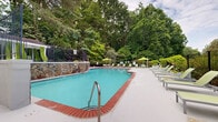 Located at 700 Summit Place Dr in Sandy Springs, Georgia, this outdoor amenity space offers a well-appointed resort-style pool area designed for relaxation and recreation. The centerpiece is a generously sized freeform swimming pool with classic brick coping and clear blue water, framed by an expansive concrete sun deck that provides ample room for lounging. Coordinated lime-green chaise lounges and patio umbrellas are arranged throughout the deck, offering both open sun exposure and shaded seating options. A striking natural stone retaining wall anchors one side of the pool, leading up to a pergola-style cabana structure accented with flowing green curtains, providing a shaded retreat for poolside gatherings. Black wrought iron fencing encloses the entire area, ensuring both security and an unobstructed view of the surrounding mature tree canopy. Raised planter beds with manicured greenery add a decorative touch to the upper terrace lounge area, which features additional seating beneath a patio umbrella. Stairways with iron railings connect the multi-level deck spaces and integrate seamlessly into the natural landscape. The dense backdrop of tall pines and hardwoods lends the space a private, tranquil atmosphere throughout.