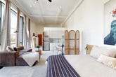 Located at 110 Somerville Ave in Chattanooga's vibrant North Shore neighborhood, this single-floor studio offers a thoughtfully designed open-plan layout that seamlessly integrates living, sleeping, dining, and kitchen areas. The space is immediately distinguished by its soaring ceilings with exposed structural detailing, lending an industrial-modern character balanced by warm, neutral tones throughout.

Floor-to-ceiling windows along one wall flood the interior with abundant natural light and frame tree-lined outdoor views, creating an airy, expansive atmosphere. Linen drapes soften the generous glazing and add a refined, residential feel. The polished concrete-style flooring flows continuously through the space, reinforcing the cohesive, contemporary aesthetic.

The kitchen area features light cabinetry, a stainless steel refrigerator, and a tiled backsplash, accompanied by a compact dining table and seating. A rattan folding screen provides a stylish visual division between the sleeping area and the rest of the studio. The sleeping area accommodates a full bed alongside a wall-mounted TV and dresser. A ceiling fan and wall-mounted mini-split unit ensure year-round comfort.

The bathroom features tile wainscoting, accessible safety bars, vinyl flooring, and a shower with a curtain enclosure.