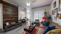 This well-appointed single-floor residence blends modern design with warm, eclectic touches throughout. The living room serves as the heart of the home, featuring light wood-look flooring, crown molding, and a ceiling fan. A striking reclaimed wood accent wall adds texture and depth, while large windows fitted with wide-slat blinds invite generous natural light. Seating is arranged around a wooden coffee table atop a vibrant area rug, complemented by a curated gallery wall that lends personality to the space. An adjacent bar-height counter provides a casual dining or workspace option. The galley-style kitchen is well-equipped with stainless steel appliances, sleek gray cabinetry, quartz countertops, and a tile backsplash, offering ample storage and counter space in an efficient layout. The dedicated dining nook features a charming bay-style corner window arrangement, bathing the round dining table in natural light. Both bedrooms are comfortably sized, each with wood-look flooring, ceiling fans, and windows providing natural light. The bedrooms are styled with mid-century modern furnishings and decorative artwork. The full bathroom features a double-sink vanity with quartz countertops, a large mirror, and a tub-shower combination, finished in a clean, contemporary palette throughout.