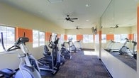 This single-floor fitness center offers a dedicated space for cardiovascular exercise, furnished with a lineup of elliptical trainers and stair-climber machines arranged along a patterned carpet floor. The room is brightened by a series of windows along two walls, allowing natural light to fill the space and providing views of the surrounding outdoor area. A distinctive orange accent stripe runs horizontally along the upper portion of the light yellow walls, adding a bold visual element that energizes the environment. Two ceiling fans are mounted overhead, promoting air circulation throughout the room and contributing to a comfortable workout atmosphere. Recessed lighting supplements the natural light for consistent illumination. A wall-mounted flat-screen television is positioned toward the far end of the room, offering entertainment during exercise sessions. A large mirror panel along one wall enhances the sense of depth and space. The overall design is clean, functional, and purpose-driven, making it a practical facility suited for regular fitness use.