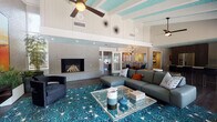 This single-floor community clubhouse showcases a striking mid-century modern aesthetic throughout, unified by a signature teal-painted ceiling with exposed white wood beams and rich dark hardwood flooring. The expansive main lounge serves as the centerpiece of the space, featuring soaring vaulted ceilings, a ceiling fan, and a floor-to-ceiling white brick fireplace. Comfortable sectional seating surrounds a glass coffee table atop a bold teal starburst-patterned area rug, creating a vibrant and inviting gathering area. Large windows and glass doors along one wall flood the lounge with natural light and offer direct views of the outdoor pool and patio. A mounted flat-screen television and colorful, large-format artwork add personality to the brick accent walls, while tall potted plants contribute a fresh, organic touch. The adjacent full kitchen is equipped with stainless steel appliances, dark wood cabinetry, a subway tile backsplash, and a generous island with bar seating — ideal for entertaining. A cozy dining nook nearby features built-in banquette seating upholstered in a geometric print, paired with a retro sputnik chandelier. The business center offers a clean, functional workspace with a wrap-around counter, track lighting, and office equipment. Throughout, the cohesive mid-century palette and curated décor create a polished, energetic ambiance.