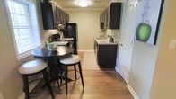 This single-floor space is defined by a well-appointed galley-style kitchen that serves as its central feature. The kitchen is outfitted with rich, dark espresso cabinetry that extends from floor to ceiling on both sides, creating a striking contrast against the warm, neutral-toned walls. Upper and lower cabinets provide generous storage, while the countertops feature a light granite-look surface that brightens the space. Black appliances, including a refrigerator, range, and microwave, complement the dark cabinetry for a cohesive, bold aesthetic. A double-basin sink with a modern faucet is neatly positioned along one wall, accompanied by a patterned runner rug that adds a touch of warmth underfoot. The warm-toned hardwood flooring runs throughout, lending a sense of continuity and a welcoming feel to the entire space. A compact dining area adjacent to the kitchen features a dark round high-top table paired with matching bar stools, making it an ideal spot for casual meals. Crown molding along the ceiling adds a refined architectural detail, while flush-mounted ceiling lighting ensures the space is well-lit. The overall ambiance is modern and functional, with a cohesive dark-and-light design palette.