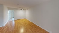 This single-story unit offers a well-organized layout with two bedrooms and one full bathroom. The living area is bright and open, featuring warm wood-style flooring that extends throughout the main spaces and a ceiling fan overhead. Large windows dressed with plantation shutters allow natural light to filter in while providing privacy, and a wall-mounted HVAC unit ensures climate control. The kitchen is efficiently designed with white cabinetry offering generous storage, tile countertops, a stainless steel sink, and black appliances including an electric range, over-the-range microwave, and dishwasher. Tile flooring grounds the kitchen area. The two bedrooms continue the wood-style flooring and maintain a clean, neutral palette with white walls and white trim. One bedroom features plantation shutters across multiple windows, flooding the space with natural light, along with a mirrored sliding-door closet. The second bedroom includes vertical blinds over a broad window. A short hallway connects the bedrooms to the full bathroom, which includes a tiled tub-shower combination, toilet, and vanity with storage. The overall ambiance is clean, light, and functional.