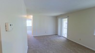 This single-floor space presents a clean, open layout centered around a spacious living area finished with wall-to-wall neutral-toned carpet and crisp white walls throughout. The room is bathed in natural light, sourced from a sliding glass door fitted with vertical blinds along one wall, offering direct access to an outdoor area and creating an airy, welcoming atmosphere. The standard ceiling height contributes to a comfortable sense of scale within the main living space. A hallway visible from the living area leads to additional rooms, suggesting a practical and straightforward floor plan. The secondary space glimpsed through the hallway also benefits from natural light via a window, maintaining a bright and consistent feel throughout. Baseboards run along the perimeter of the room, adding a clean finishing detail to the interior. The neutral color palette and simple finishes provide a versatile backdrop suitable for a variety of interior styles. Overall, the space is well-lit, open, and move-in ready, with a cohesive design that prioritizes functionality and ease of living.