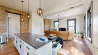 Located at 102 Main St in Hamilton, Ohio, this single-floor, one-bedroom, one-bathroom unit offers a well-designed layout that balances industrial character with contemporary finishes. The open-plan kitchen and living area serves as the heart of the space, where an exposed brick accent wall creates a striking focal point against clean white walls and light wood-look flooring that runs throughout. The kitchen features white shaker cabinetry, dark granite-style countertops, a subway tile backsplash, stainless steel appliances, and a peninsula with pendant lighting overhead—ideal for both cooking and casual dining. The living area is generously proportioned, with multiple large double-hung windows framed in dark trim that flood the room with natural light and offer outdoor views. A ceiling fan adds a functional accent overhead, while exposed ductwork contributes to the industrial aesthetic. The spacious primary bedroom continues the consistent flooring and neutral palette, with a ceiling fan and a window providing natural light. The full bathroom features wood-look flooring, white cabinetry with dark countertops, and a shower-tub combination. A dedicated closet completes the layout, providing practical storage within this well-appointed unit.