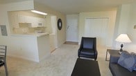 This single-floor space showcases an open-concept design that seamlessly connects the kitchen and living area, creating a cohesive and functional layout. The living area features light-toned carpeting that lends warmth underfoot, while the soft, neutral cream walls contribute to a bright and airy ambiance throughout the space. A closet with bifold doors provides convenient built-in storage.

The kitchen is positioned adjacent to the living space, separated by a peninsula-style counter that serves as both a workspace and a casual dining surface. White cabinetry lines the kitchen walls, complemented by a white gas range and a built-in microwave overhead. The kitchen floor transitions to a light-toned hard surface, providing a practical contrast to the carpeted living area. Recessed fluorescent lighting above the kitchen counter ensures a well-lit workspace.

The entryway, visible from the living area, features a tile floor transition and a white front door that anchors the layout. A decorative circular mirror mounted near the entryway adds a subtle design accent. Overall, the space maintains a clean, understated aesthetic with a practical, open feel.