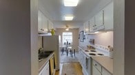 This single-story, 2-bedroom, 2-bathroom home in Monterey is designed with practicality and comfort in mind. The galley-style kitchen is a standout feature, offering ample white cabinetry on both sides for generous storage, a double basin sink, and a full suite of white appliances including an electric range with overhead ventilation. Flush-mounted ceiling lighting illuminates the kitchen workspace efficiently, while a natural woven rug adds a warm, textured touch to the hardwood-style flooring beneath. The kitchen flows seamlessly into a bright dining area, where a ceiling fan with a light fixture helps maintain a comfortable atmosphere. A window allows natural light to fill the space, creating an open and airy feel. The dining area accommodates a bistro-style table and chairs, making it an inviting spot for everyday meals. The textured walls throughout add subtle visual depth to the interior. The overall ambiance of this residence is clean, functional, and welcoming, with a neutral color palette that provides a versatile backdrop for personalization. The thoughtful layout makes efficient use of space, ensuring both bedrooms and bathrooms are comfortably integrated into the single-level floor plan.