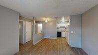 This single-floor unit offers a well-organized layout that balances functionality and comfort. The open-plan living area flows directly into the kitchen, creating a connected, airy feel. Warm hardwood-style flooring runs throughout the main living space, complemented by neutral gray walls and a textured ceiling. A ceiling fan provides additional comfort to the living area, which benefits from natural light filtering in through windows.

The kitchen is efficiently designed with ample white cabinetry providing generous storage, speckled laminate countertops, a stainless steel sink, a black dishwasher, and a white refrigerator. A pass-through opening connects the kitchen to the living space, enhancing the open feel.

The two bedrooms feature plush carpeting, gray walls, and windows with outdoor views, each equipped with a wall-mounted HVAC unit for individual climate control. One bedroom also includes a built-in desk or shelf, adding a practical workspace element.

The bathroom is appointed with marble-patterned wall surround tiles encasing a full soaking tub and shower combo, a white vanity with speckled countertop, and classic fixtures. The overall ambiance of the unit is clean, neutral, and move-in ready, offering a comfortable and practical living environment.
