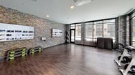 This single-floor fitness studio is thoughtfully designed to support a variety of workout styles in a visually dynamic environment. The space is defined by its striking mosaic-patterned accent walls, which add an energetic, artistic character throughout. Rich, dark hardwood-style flooring grounds the space and provides a durable, clean surface ideal for group exercise and individual training alike.

The studio is divided into distinct functional zones. One area is dedicated to indoor cycling, featuring a row of stationary spin bikes positioned directly in front of expansive floor-to-ceiling windows. These large, black-framed windows flood the space with natural light and offer open views of the surrounding urban streetscape, creating an engaging backdrop during workouts.

An adjacent open-floor exercise area provides ample room for stretching, yoga, barre, and other group fitness activities. This zone features mounted flat-screen displays for on-demand workout content, a ballet barre along the window wall, and stability balls for functional training. Step platforms are neatly stored along the accent wall, keeping the floor unobstructed.

The studio connects seamlessly to additional amenity spaces through two access points set against a deep teal wall — one leading to what appears to be a main fitness equipment room, and the other to a separate amenity area. Recessed lighting complements the abundant natural light, ensuring the space remains bright and inviting at all times.