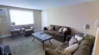 This single-floor space offers a warm and functional layout designed for comfortable everyday living. The living room serves as the central gathering area, featuring a neutral color palette of soft beige and warm gray tones that create a welcoming ambiance throughout. The room is furnished with a generously sized tufted sofa, a matching loveseat, and an accent armchair, all upholstered in rich dark brown fabric and accented with decorative patterned pillows. A rectangular tile-top coffee table with a wrought iron base anchors the seating arrangement, adding a touch of understated style. Carpeted flooring runs throughout, contributing to the room's cozy and relaxed feel. A large window allows natural light to fill the space, brightening the interior and enhancing its open atmosphere. The clean white walls and simple, uncluttered design give the space a practical yet inviting character. A white interior door provides access to additional areas of the home, completing a layout that balances simplicity with comfort.