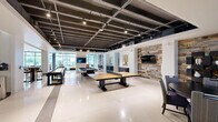 Located at 3700 Ross Avenue in Dallas, TX, this spacious single-floor amenity lounge offers an impressive and versatile entertainment environment designed in a contemporary industrial-modern style. The exposed structural ceiling with painted steel beams and integrated track lighting creates a distinctive loft-like aesthetic throughout the generously proportioned open floor plan.

The space is thoughtfully divided into several distinct activity and lounge zones. A dedicated golf simulator room features a large projection screen, turf hitting mat, and a sleek club storage console set against a sculptural textured accent wall, offering an immersive experience. Adjacent to this area, a full-size billiards table serves as a central focal point, surrounded by polished terrazzo flooring that extends throughout the space.

Multiple seating arrangements cater to various social settings, including clusters of rounded cognac leather lounge chairs with a live-edge wood coffee table, tufted blue velvet wingback chairs, and upholstered sofas. A long communal bar-height counter with pendant lighting and mounted flat-screen televisions provides a lively social hub. A shuffleboard table and additional game areas further enhance the recreational offerings.

Floor-to-ceiling windows along the perimeter flood the interior with natural light while framing views of the surrounding Dallas cityscape. Curated artwork displayed on crisp white walls adds cultural sophistication to this dynamic, amenity-rich environment.