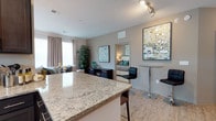 This single-story, 2-bedroom, 2-bathroom residence is designed with an open-concept floor plan that promotes a seamless flow between the kitchen, dining, and living areas. The kitchen features a large granite-topped island with bar seating, providing both a functional workspace and an ideal spot for casual dining or entertaining. Light wood-tone flooring extends throughout the main living areas, contributing to a warm, cohesive aesthetic.

The living area is generously sized and benefits from natural light streaming through large windows dressed with neutral curtains. A dedicated dining nook with a high-top table and bar stools adds a versatile space for meals or work. Neutral-toned walls serve as a clean backdrop, complemented by contemporary artwork and decorative accents.

The bedroom, visible from the main living area, is well-proportioned and features coordinated decor with teal accent pillows and warm lighting. Recessed track lighting in the kitchen provides targeted illumination, while the overall layout maximizes functionality. The space balances modern design elements with a comfortable, livable ambiance suited for everyday use.