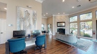 This single-floor space is thoughtfully designed to balance elegance with functionality. Soaring ceilings with crown molding lend a grand sense of scale throughout, while rich, wide-plank hardwood flooring grounds the interior with warmth and continuity. Floor-to-ceiling windows and oversized glass entry doors flood the main gathering area with natural light, framing views of mature trees and lush landscaping beyond.

The central lounge area anchors the space with a contemporary sofa arrangement set atop a layered area rug, positioned in front of a striking corner fireplace clad in textured whitewashed stone and topped with a framed mirror. A sputnik-style chandelier overhead adds a refined, modern touch to the open entry zone.

A dedicated workstation area features dual monitors set against a dramatic oversized abstract artwork, with teal ergonomic chairs providing a bold accent. A small conference or meeting nook with a round wood table and navy chairs sits bathed in natural light from surrounding windows, with curated artwork adorning the walls.

A quieter alcove offers two wingback chairs beside a digital kiosk, creating an intimate consultation or waiting area. Decorative copper wall medallions and high-mounted art installations contribute to the space's polished, curated aesthetic throughout.