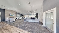 Located at 3580 E Alexander Rd in Las Vegas, NV, this single-floor leasing office and community lounge space combines contemporary design with welcoming functionality. The open-plan layout is anchored by a striking marble-clad reception desk, accented by a dramatic full-height marble feature wall that commands immediate attention upon entry. Soaring vaulted ceilings with recessed lighting and a sculptural multi-arm pendant chandelier lend an airy, upscale ambiance throughout the space.

The flooring transitions seamlessly between rich wood-look planks near the entry and deeply patterned carpeting throughout the main lounge and reception areas. A dedicated seating lounge features elegant white wingback chairs arranged around glass-top tables, centered on a tiled accent wall with a mounted flat-screen display and a sleek linear fireplace below. Opposite, a row of workstations provides a functional leasing area. Decorative wall art and curated greenery add warmth and personality throughout.

Large floor-to-ceiling windows and glass doors flood the interior with natural light while framing direct views of an outdoor pool area lined with palm trees, blurring the boundary between indoor and outdoor living. A casual dining nook with a round marble-top table and upholstered chairs completes the inviting, resort-inspired atmosphere of this thoughtfully designed community hub.