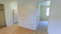 This single-story space is thoughtfully arranged across distinct zones visible from a central transitional area. The kitchen section features light-toned hardwood flooring, white walls, and a full-size white refrigerator, contributing to a clean and bright atmosphere. A partition wall separates the kitchen from the adjoining room, creating a defined sense of separation between spaces while maintaining an open feel overall.

Through the doorway, a carpeted room is visible, finished in a neutral, light gray-beige tone that adds warmth underfoot. This area benefits from natural light entering through a window on the far wall, brightening the space and enhancing its open character. The walls throughout are painted in soft white and light neutral tones, contributing to a cohesive and airy aesthetic.

The textured ceiling is consistent across both visible areas, and standard electrical outlets and light switches are positioned at convenient heights throughout. The transition between the hardwood and carpeted zones is clean and well-defined, reflecting a practical and straightforward interior layout suited for everyday living.