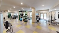 This fitness center offers a comprehensive workout environment designed to accommodate a variety of exercise routines. The space is defined by its open, single-floor layout and bright, welcoming atmosphere, enhanced by recessed lighting and large windows and glass double doors that invite generous natural light throughout the area.

The flooring features a distinctive yellow and grey checkerboard pattern, adding a clean, energetic aesthetic to the space. Strength training is well-supported with a dedicated free weights section, including a full dumbbell rack, a multi-functional green cable and pull-up rig, and an assortment of medicine balls and stability equipment. Barbell and plate weight stations are also available for more intensive strength work.

The cardio section is equipped with multiple treadmills and other aerobic machines, complemented by a wall-mounted flat-screen television — ideal for staying entertained during longer sessions. Support columns are thoughtfully integrated into the layout without disrupting the flow of the space.

Neutral-toned walls and a well-organized arrangement of equipment contribute to an orderly, motivating environment. The fitness center is well-suited for both casual users and dedicated athletes looking for a fully functional, amenity-rich workout space.