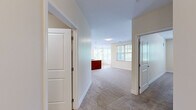 This single-floor residence offers a thoughtfully designed layout with a clean, neutral color palette and a variety of flooring materials suited to each space. The kitchen serves as a functional centerpiece, featuring rich cherry wood cabinetry, granite countertops, stainless steel appliances including a range, microwave, and refrigerator, and a peninsula island with additional counter space. Warm hardwood-style flooring flows through the kitchen and into the adjacent dining area, which is bathed in natural light through a large corner window arrangement and accented by an elegant chandelier overhead. The living area is generously proportioned, finished with plush neutral carpeting and connected to a central hallway that provides easy access to the rest of the home. Multiple bedrooms offer comfortable carpeted floors and ample natural light through windows overlooking lush outdoor greenery. One bedroom provides convenient access to an in-unit laundry area. The full bathroom is finished with tile flooring, a cherry wood vanity with granite countertop, and a full tub-shower combination with an accessibility grab bar. Throughout the residence, crisp white trim, panel doors, and softly painted walls create a bright, cohesive atmosphere.