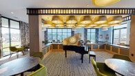 Located at Riverwalk in Plainsboro, NJ, this single-floor community lounge is a thoughtfully designed social space that blends mid-century modern aesthetics with contemporary comfort. The layout is generous and open, flowing seamlessly between distinct functional zones that together create a cohesive, inviting atmosphere.

At the heart of the space sits a grand piano, serving as a striking focal point beneath a dramatic cluster of large, gold drum pendant lights that cast a warm, ambient glow throughout the room. The bold black-and-white geometric patterned carpet anchors the lounge seating area, where olive green upholstered chairs and dark marble-topped tables are arranged to encourage both intimate conversation and relaxed gatherings.

A full-service bar runs along one side of the space, featuring a sleek dark countertop, brass tap fixtures, and an architectural brass shelving display set against white tile, offering a polished, sophisticated touch. Bar-height wooden stools line the counter for casual seating.

Floor-to-ceiling windows along the perimeter flood the interior with natural light and frame views of the surrounding Riverwalk neighborhood. Framed landscape artwork adorns the walls, complementing the warm wood paneling and light-toned architectural millwork. A glassed private area provides a more enclosed setting within the broader lounge, adding versatility to the overall space.