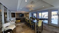 This sophisticated single-floor social space is designed with both comfort and style in mind. The open-concept layout seamlessly connects a dining and lounge area, creating an inviting environment for gatherings and entertainment. At the heart of the space, dark espresso-toned bar-height tables are paired with green upholstered chairs, offering a contemporary and elegant seating arrangement. The lounge area features plush seating positioned to face a wall-mounted flat-screen television, framed by a striking textured stone accent wall that adds depth and visual interest. A wet bar or kitchenette area anchors the entertainment zone, providing added functionality for hosting. The ceiling features a combination of recessed lighting and a prominent drum pendant fixture, casting warm, layered illumination throughout. Large floor-to-ceiling windows and glass doors flood the interior with natural light while offering views of an outdoor area, enhancing the sense of openness. The flooring consists of large-format stone or tile in warm neutral tones, contributing to the upscale aesthetic. Overall, the space exudes a polished, modern ambiance suited for both casual socializing and more formal gatherings.