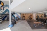 Located at 318 W Wilson Ave in Glendale, California, this two-story fitness center is a well-appointed facility designed to support a full range of workout routines. The ground floor welcomes visitors with soaring double-height ceilings and expansive floor-to-ceiling windows that flood the space with natural light. This level houses functional training equipment including a multi-station cable machine, a heavy punching bag, medicine ball racks, a BOSU balance trainer, and a wall-mounted display for interactive fitness programming. A staircase with sleek metal railings connects the two floors, creating an open, airy atmosphere between levels. The upper floor provides a spacious cardio and strength training area finished with neutral carpet tile flooring. It features a row of Cybex elliptical trainers, upright and recumbent stationary bikes, treadmills, and a rowing machine, offering diverse options for cardiovascular conditioning. A dedicated stretching and floor exercise area with workout mats rounds out the upper level. Throughout both floors, bold blue and white diagonal graphic murals energize the aesthetic, complemented by framed Los Angeles Lakers and Dodgers jerseys that reinforce a spirited, sports-inspired theme. The overall ambiance is modern, clean, and motivating, making this a comprehensive fitness destination.