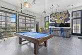 Located at 2315 South 11th Avenue in Bozeman, Montana, this single-floor game room delivers a lively, mountain-inspired atmosphere with a distinctly urban edge. The open-plan layout makes excellent use of its generous footprint, with polished concrete floors and elevated ceilings that amplify the sense of space throughout.

Floor-to-ceiling glass garage doors line one wall, flooding the interior with abundant natural light and offering views of the surrounding neighborhood. Industrial-style pendant lights hang from exposed ceiling infrastructure, reinforcing the room's modern, utilitarian aesthetic.

A central pool table with a bold blue felt surface serves as the main focal point, complemented by a rustic wood-paneled bar counter with a dark granite top and colorful metal barstools in yellow and blue. A dedicated kitchenette area features dark wood cabinetry, a granite countertop, a mounted TV, and a striking hand-lettered "Game On" mural that commands attention.

A mountain-themed art installation with a "Ski Repair" sign anchors one corner, nodding to Bozeman's outdoor recreation culture and adding a locally inspired decorative element. The space also includes additional seating along the glass doors, creating an ideal area for socializing. Overall, this game room combines playful design, functional amenities, and regional character into a cohesive and energetic communal space.