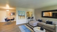 This single-floor residence offers a thoughtfully designed open-concept layout that maximizes both space and functionality. The living area serves as the heart of the home, featuring rich hardwood-style flooring that extends seamlessly throughout, lending warmth and cohesion to the interior. Neutral-toned walls provide a versatile backdrop, complemented by carefully chosen artwork that adds character and color to the space.

A standout feature is the pass-through kitchen bar, which connects the kitchen to the living area, creating an inviting space for casual dining or entertaining. Bar-height seating flanks the counter, making it ideal for conversation while meals are prepared. The kitchen beyond is equipped with stainless steel appliances, visible through the pass-through opening.

The living room is generously proportioned, accommodating a large sectional sofa, an ottoman, and a round accent coffee table with ease. A striped area rug in blue, grey, and green tones anchors the seating arrangement, adding visual interest underfoot. A large framed mirror on the wall enhances the sense of openness. Adjacent to the main living space, a separate dining area is also visible, offering additional flexibility for everyday living. The overall ambiance is modern, clean, and welcoming.