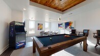 Located at 4000 Revere Blvd in Sherrills Ford, NC, this single-floor recreational space combines contemporary design with a welcoming, social atmosphere. The centerpiece of the room is a solid wood-framed pool table with a dark felt surface, positioned beneath a striking tray ceiling clad in warm, natural wood planking and illuminated by a sleek circular pendant light. Dark wood-toned tile flooring anchors the space, complemented by area rugs that define distinct activity zones. A wall-mounted flat-screen television and a classic upright arcade game add to the entertainment options, making the space well-equipped for recreation. A dedicated lounge area features a set of plush, curved swivel chairs arranged on carpet, bathed in natural light streaming through large windows that offer open outdoor views of the surrounding landscape. Vibrant, large-scale abstract paintings displayed throughout the room inject bold color and artistic energy against the crisp white walls. Recessed lighting supplements the natural light, ensuring the space is well-lit throughout the day. Bar-height seating along one wall provides additional gathering spots. The overall ambiance is modern yet inviting, striking a balance between lively entertainment and relaxed socializing.