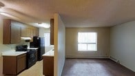This single-floor space is organized around an open-concept design that connects the kitchen and living area, creating a functional and cohesive layout. The living area features wall-to-wall carpet, neutral-toned walls, and a textured ceiling, lending a straightforward and practical character to the space. Baseboard heaters line the walls, providing efficient heating throughout. Natural light enters through windows fitted with vertical blinds, brightening the interior.

The kitchen is partially separated from the living area by a half-wall partition, which also serves as a breakfast bar with a light-colored countertop. The kitchen is equipped with black appliances, including a refrigerator and stove, complemented by warm wood-toned cabinetry. An overhead fluorescent light fixture provides task lighting over the workspace. The kitchen floor transitions to a hard surface, offering a practical contrast to the carpeted living area.

Overall, the space presents a clean, utilitarian layout well-suited for everyday living, with clearly defined zones for cooking and relaxing while maintaining an open, connected feel.